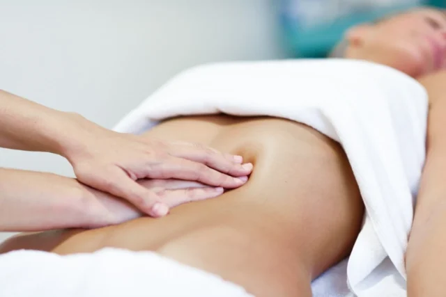 Persona tumbada boca arriba cubierta con una toalla blanca mientras recibe un masaje de drenaje linfático abdominal. Las manos del masajista presionan suavemente la zona del abdomen, sugiriendo una sesión de relajación o un tratamiento terapéutico. El fondo se difumina para centrar la atención en el masaje.
